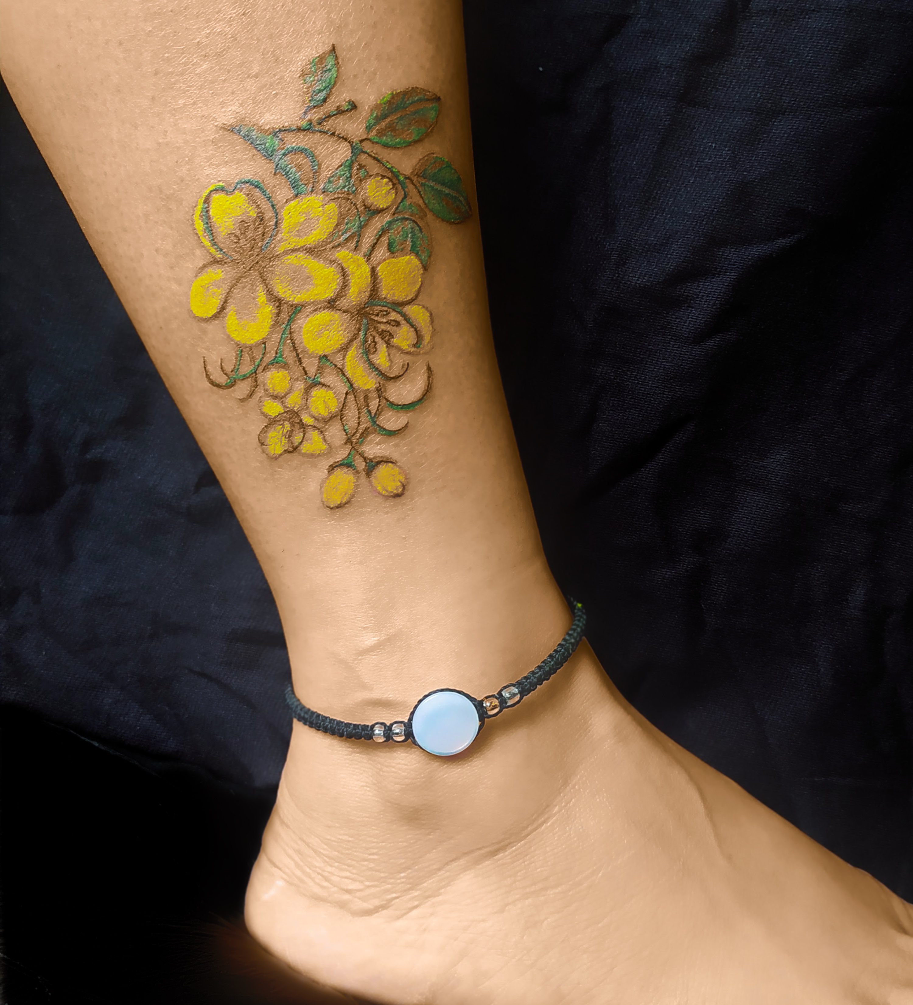 Tatuagem feminina nas pernas de flores amarelas no tornozelo, estilo colorido e delicado, feita pelo tatuador feminino Cris Santos em Mogi das Cruzes e região.